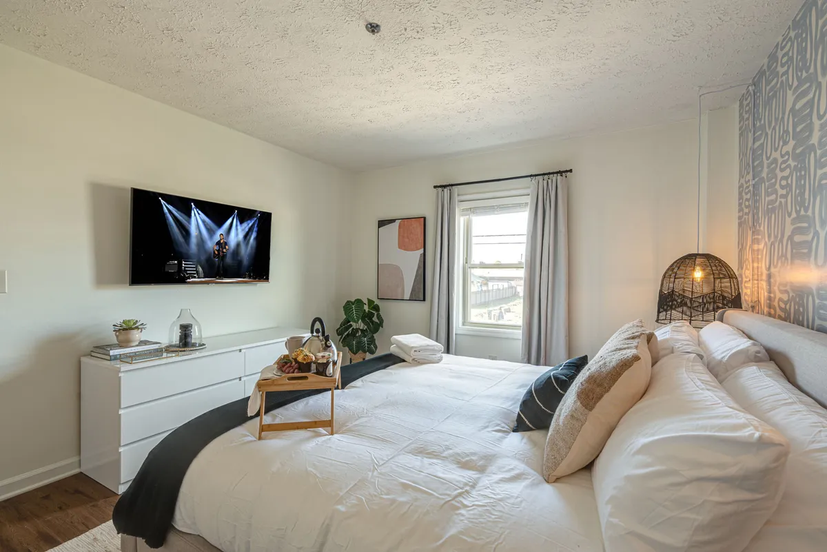 A cozy, modern bedroom featuring a well-made bed with decorative pillows, a smart TV mounted on the wall, a window with curtains allowing natural light, stylish artwork, and a decorative lamp on the side table.