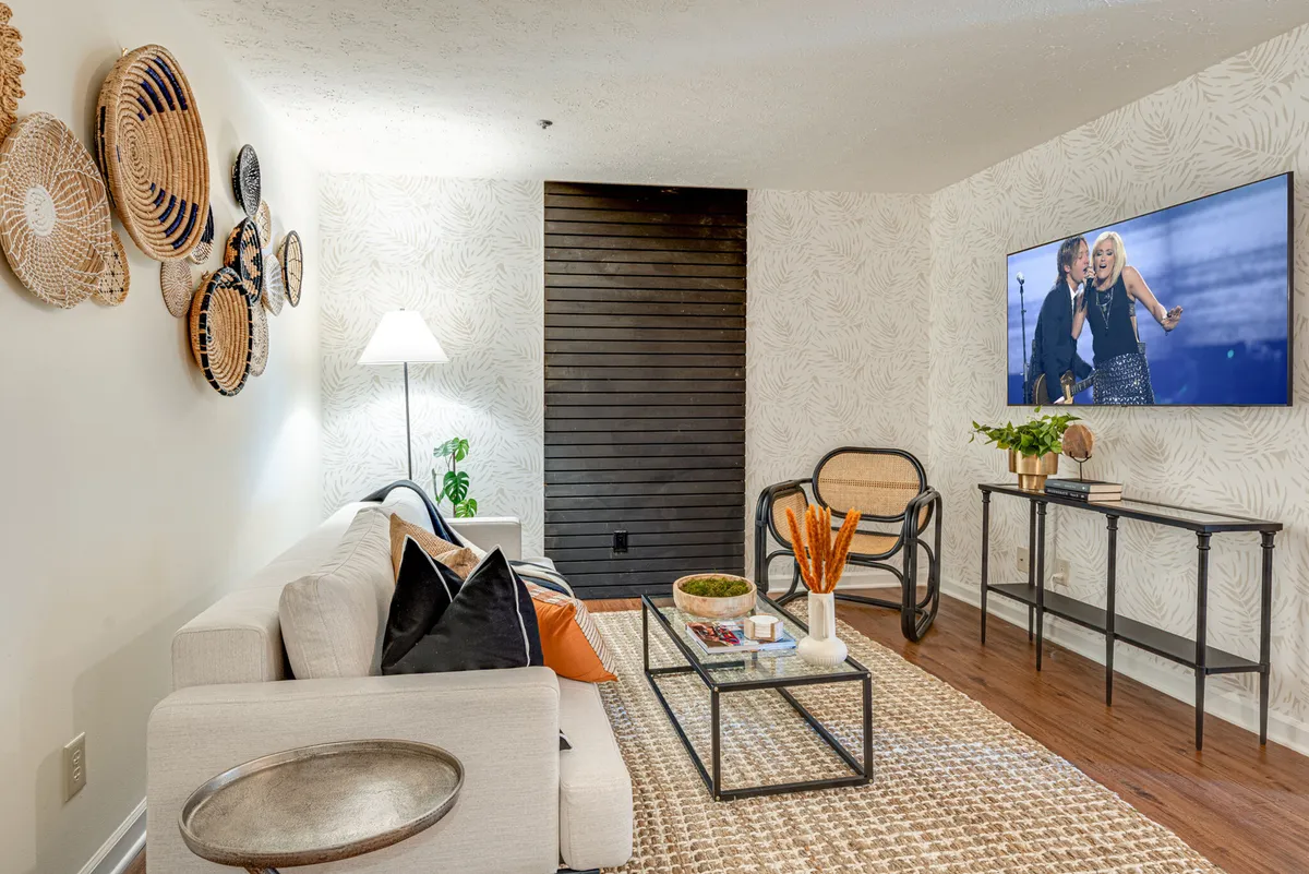 The image features a bright, inviting living room with a plush sofa adorned with colorful pillows, woven boho wall art, a modern TV displaying entertainment content, a vibrant decorative plant, and a stylish coffee table with curated décor. The space is filled with natural light, creating a chic and cozy atmosphere perfect for relaxing or socializing.