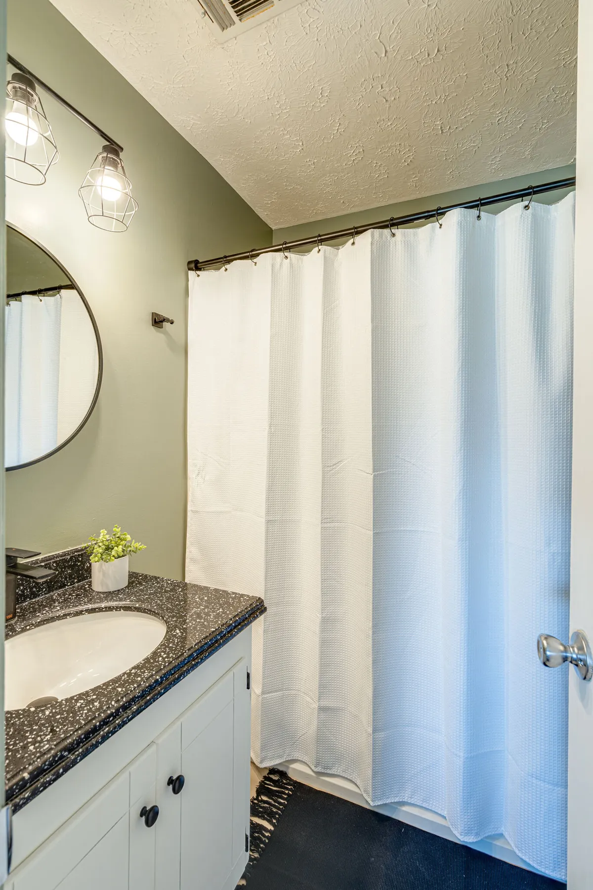 A calm, refreshing bathroom with a round mirror above a dark granite countertop, a white shower curtain, green painted walls, and a small potted plant adding a touch of nature.