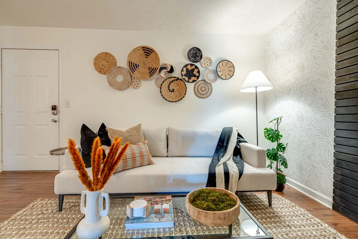 A bright living area featuring woven wall art, a comfortable sofa, a coffee table with decorative accents, and a lush indoor plant. The environment is cozy and stylish, inviting guests to relax, socialize, or unwind with loved ones—including pets.
