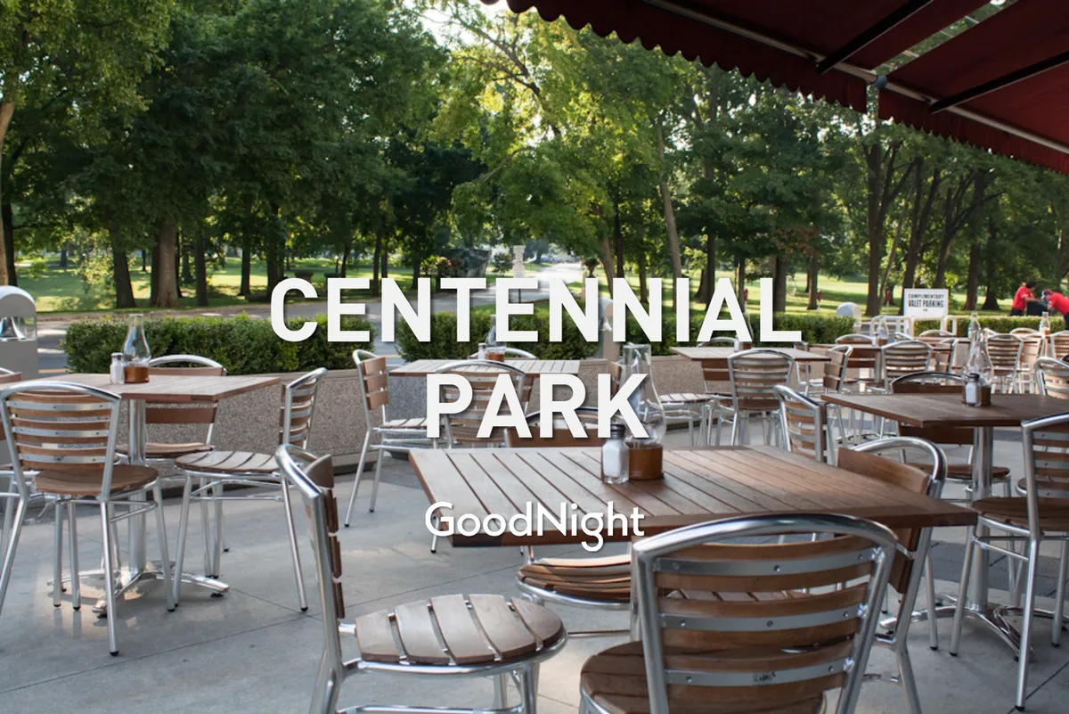 A lively outdoor dining area with wooden tables and metal chairs, surrounded by lush greenery and a park pathway. The setting is casual and friendly, suggesting opportunities for dining, socializing, and enjoying local cuisine outdoors.